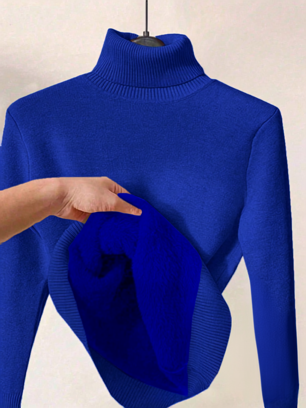 A bright royal blue ribbed turtleneck sweater displayed on a hanger, with a hand holding up a folded section of the blue knit fabric to show texture and drape.