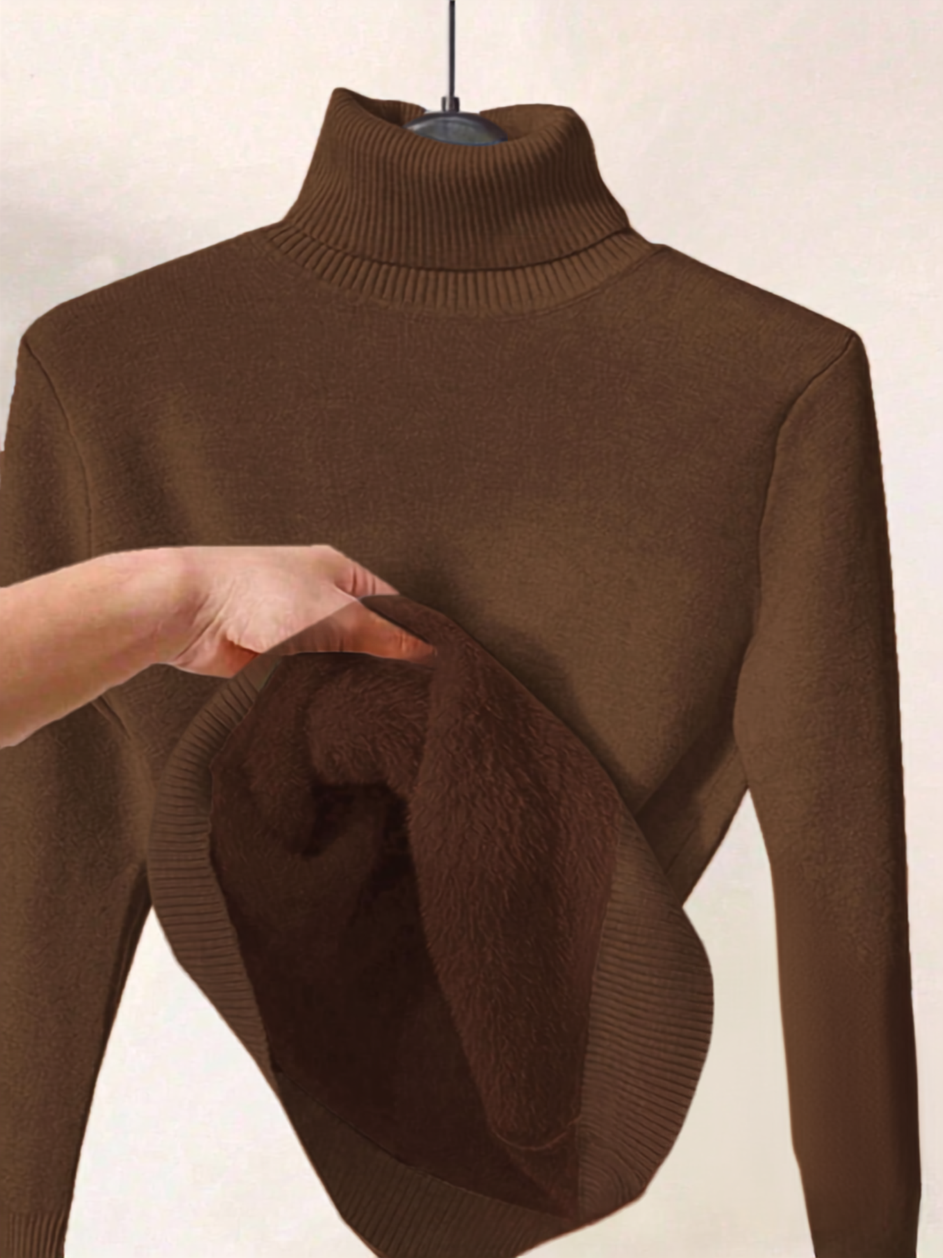 A brown ribbed turtleneck sweater displayed on a hanger, with a hand lifting the hem to reveal a plush, soft inner lining.