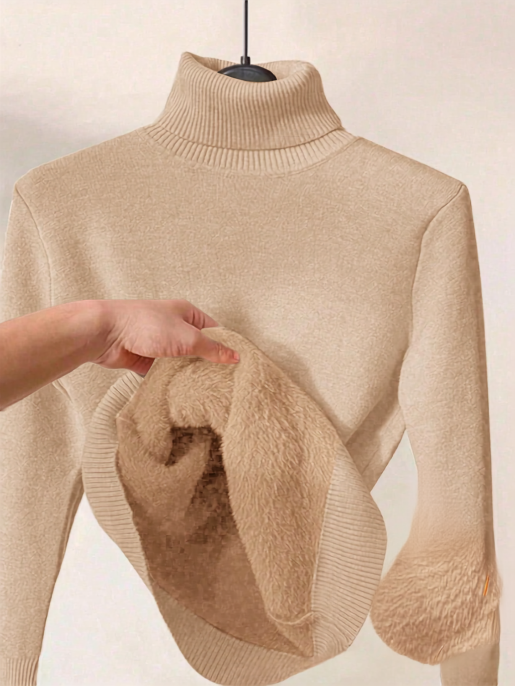 A beige ribbed turtleneck sweater displayed on a hanger, with a hand pulling out its thick, plush fleece lining to show the soft texture.