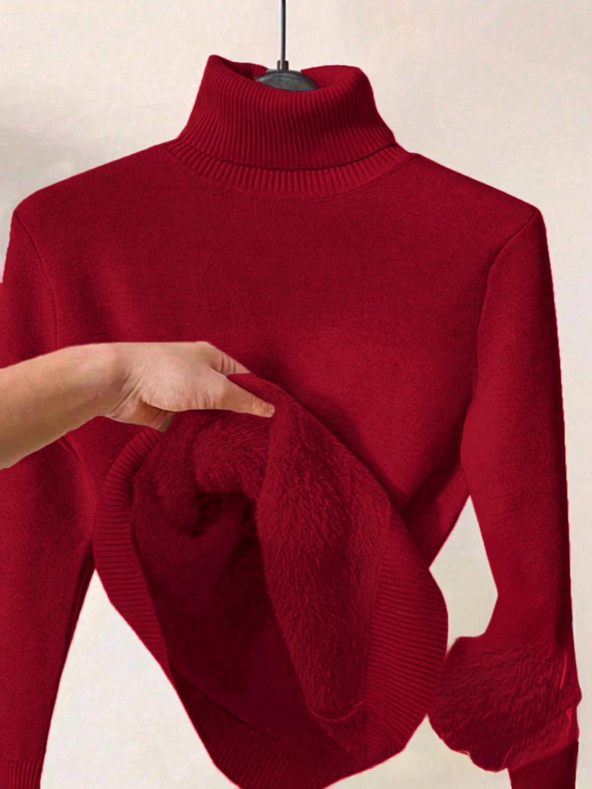 A deep red ribbed turtleneck sweater displayed on a hanger, with a hand pulling out the soft plush lining of the collar to show the inner texture.