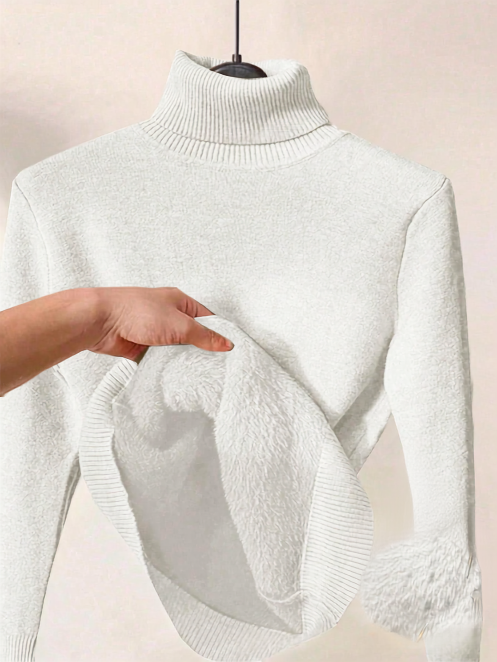 A white ribbed turtleneck sweater displayed on a hanger, with a hand lifting the hem to show a soft, plush lining.
