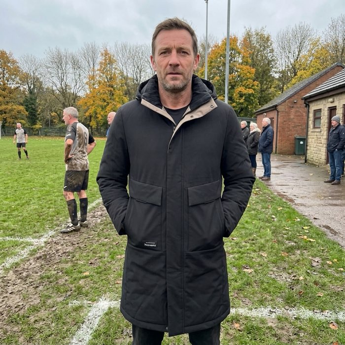 Man in a long hooded puffer coat beside a football pitch